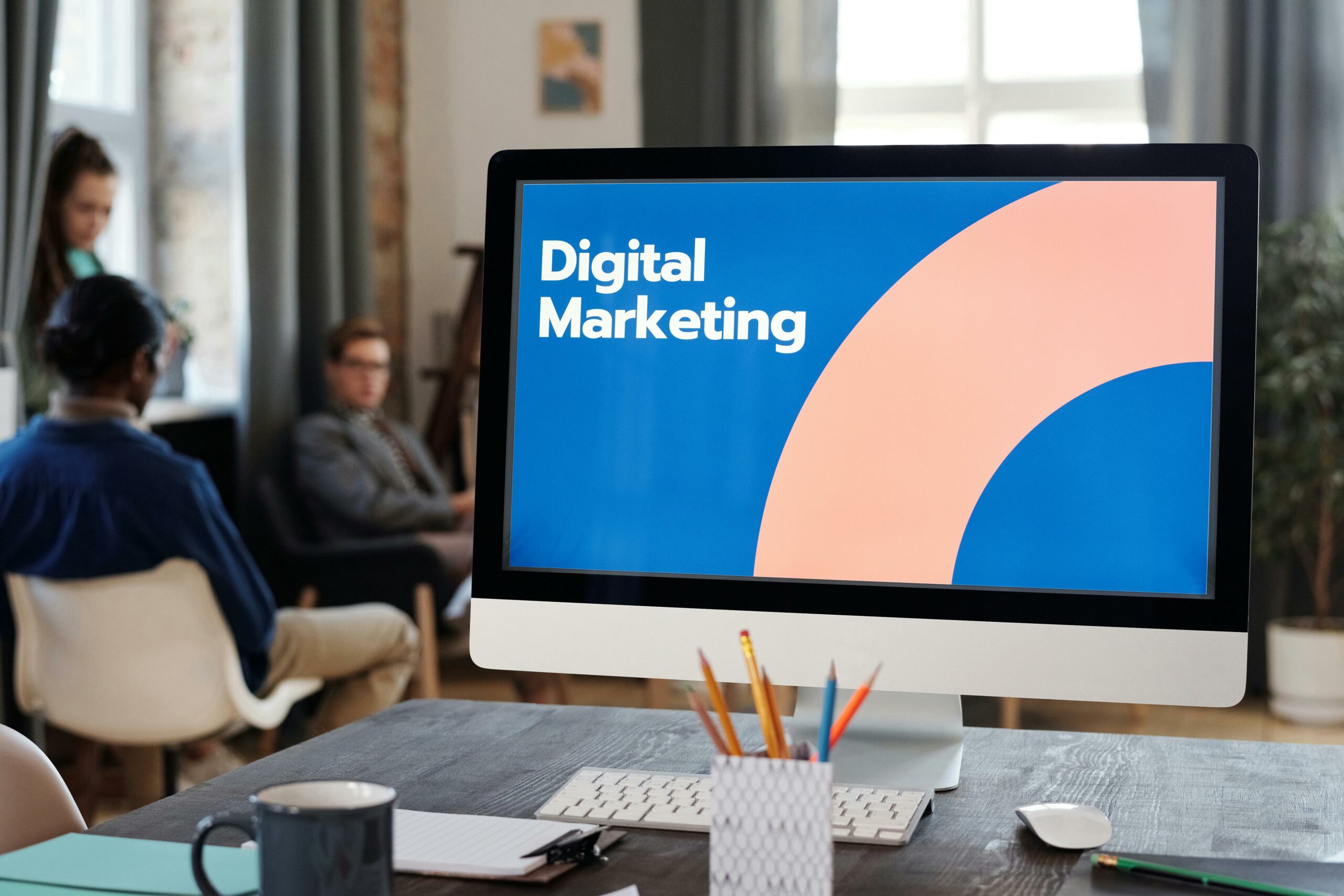 Computer screen on a desk displaying 'Digital Marketing,' with a blurred background featuring three individuals – two seated on chairs and one standing. Shallow depth of field emphasizes the focal point on the screen and workspace.