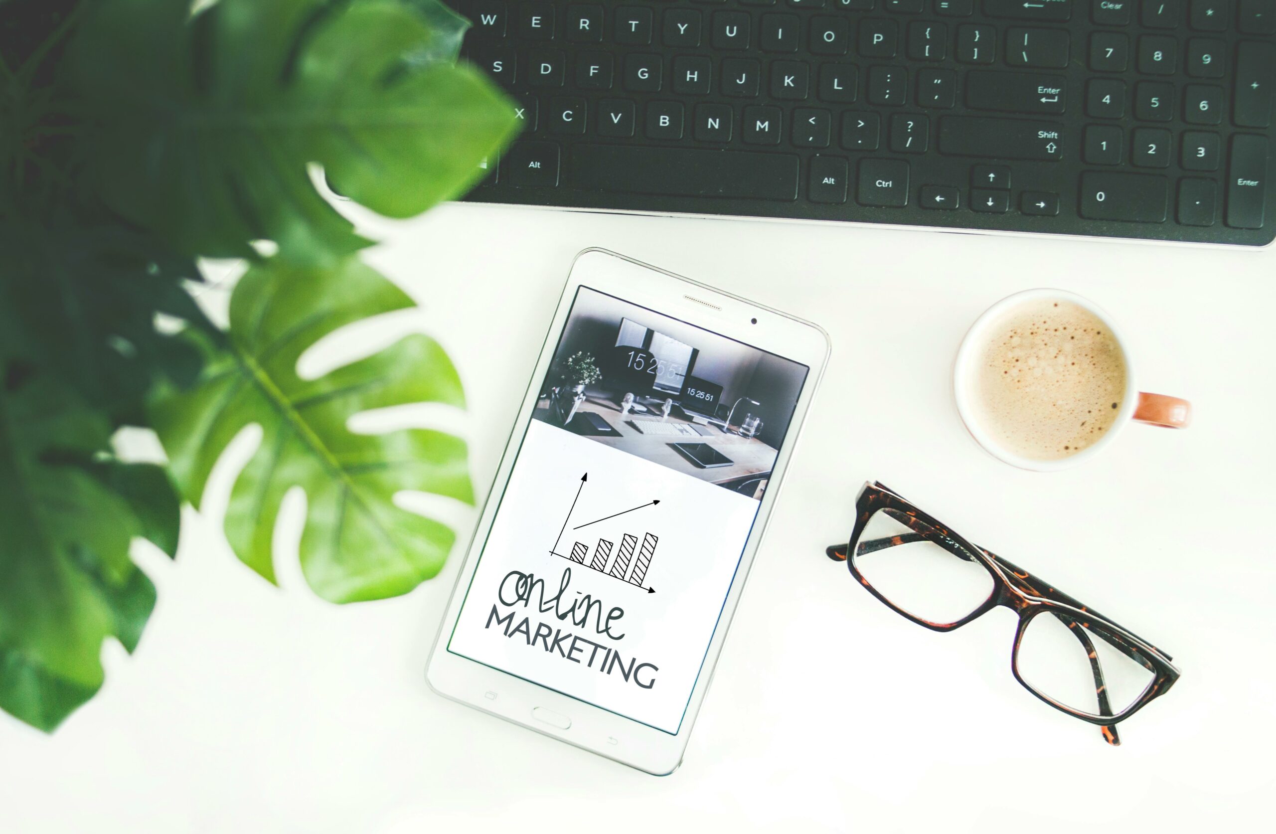 Phone displaying a growth graph with the label 'Online Marketing.' Accompanied by reading glasses, a cup of coffee, a computer keyboard, and a plant, the image portrays a workspace with digital tools for strategic online marketing.